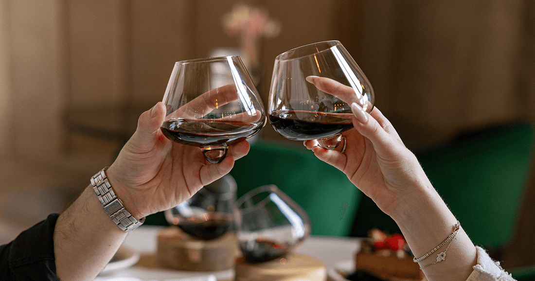 Two people toasting with Aequilibrium Wine Glasses by Audacem, filled with red wine, in an elegant indoor setting.