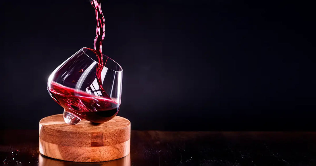 Red wine being poured into a tilted, hand-blown wine glass on a wooden coaster against a dark background.