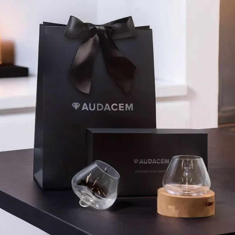 Audacem gift set featuring Aequilibrium glasses, black gift box, and ribboned bag displayed on a modern dark table.