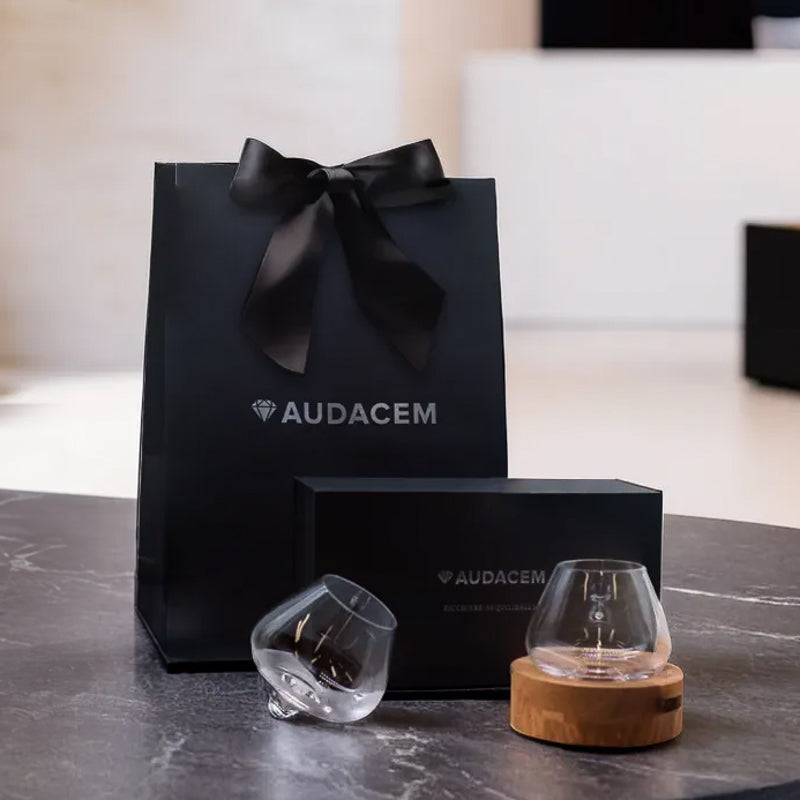 Audacem Aequilibrium gift set with two glasses, black box, and branded gift bag on a dark stone table.