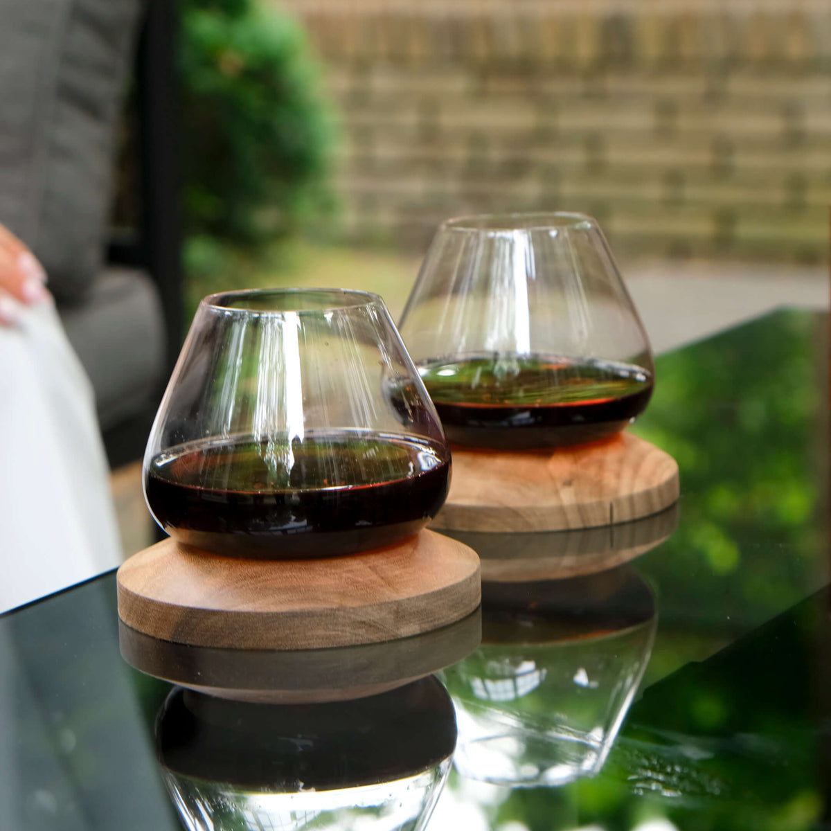 Aequilibrium Wine Glasses by Audacem with dark red wine on matching wooden coasters, displayed on reflective outdoor table for an elegant drinking experience