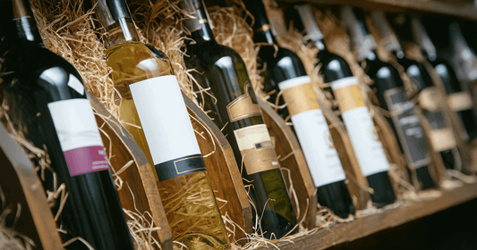 Assorted bottles of wine with blank and artistic labels displayed on a wooden shelf lined with straw.