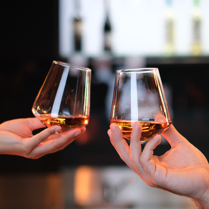 Two hands toasting with Aequilibrium Universal Glasses filled with amber liquor.