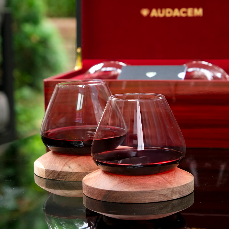 Two Aequilibrium wine glasses with red wine on wooden coasters, presented in front of the Audacem gift box on a reflective outdoor table.