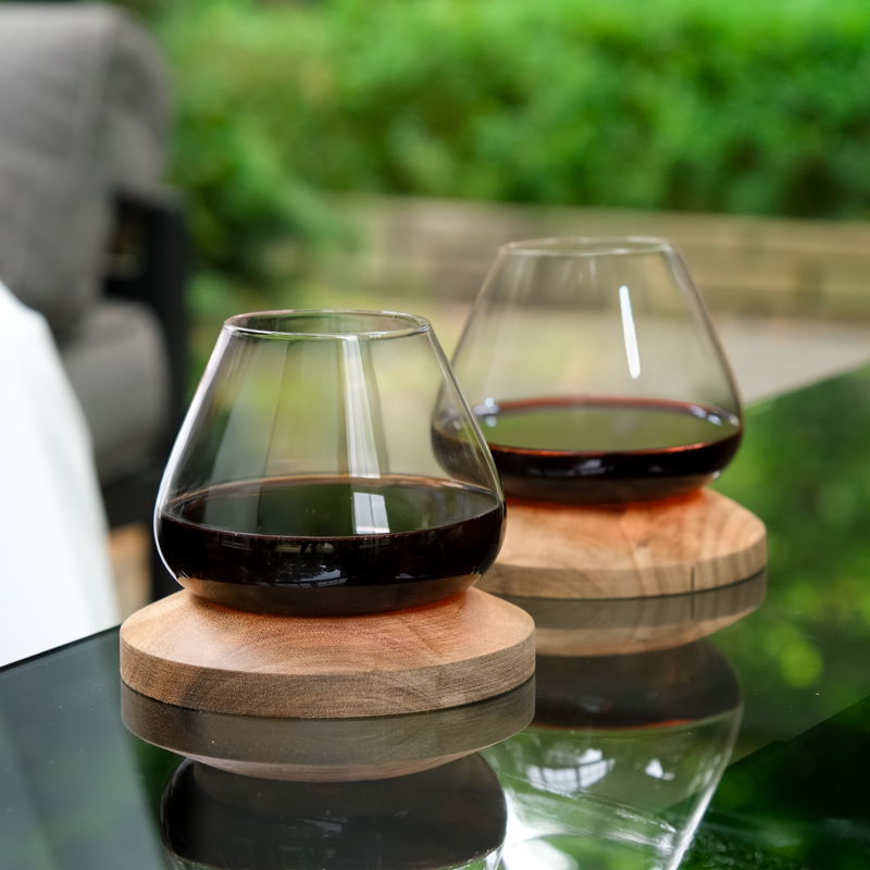 Two Aequilibrium red wine glasses on handcrafted wooden coasters, placed on outdoor glass table with garden background.