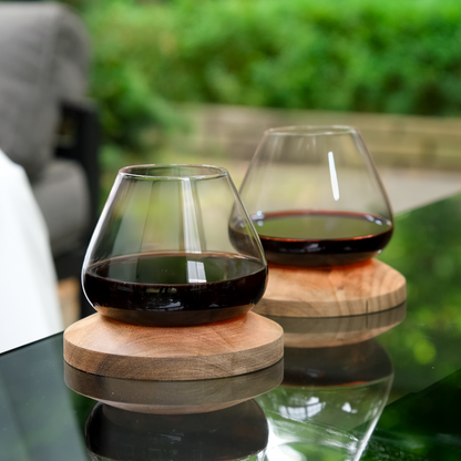 Two Aequilibrium red wine glasses on handcrafted wooden coasters, placed on outdoor glass table with garden background.