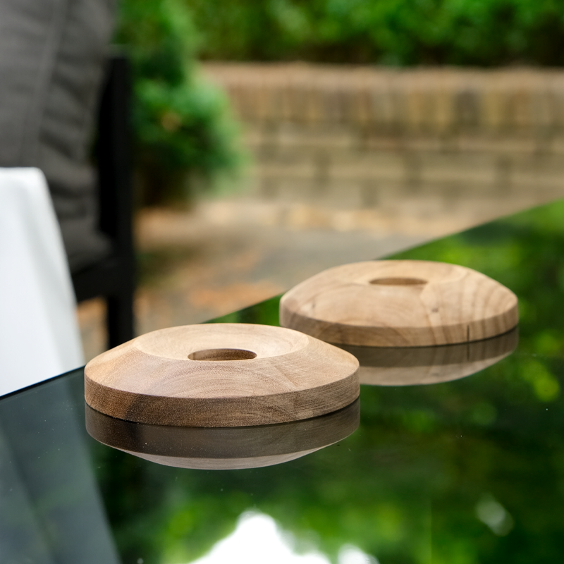 Two handcrafted wooden coasters with central indent, displayed on a reflective glass table in an outdoor garden setting.