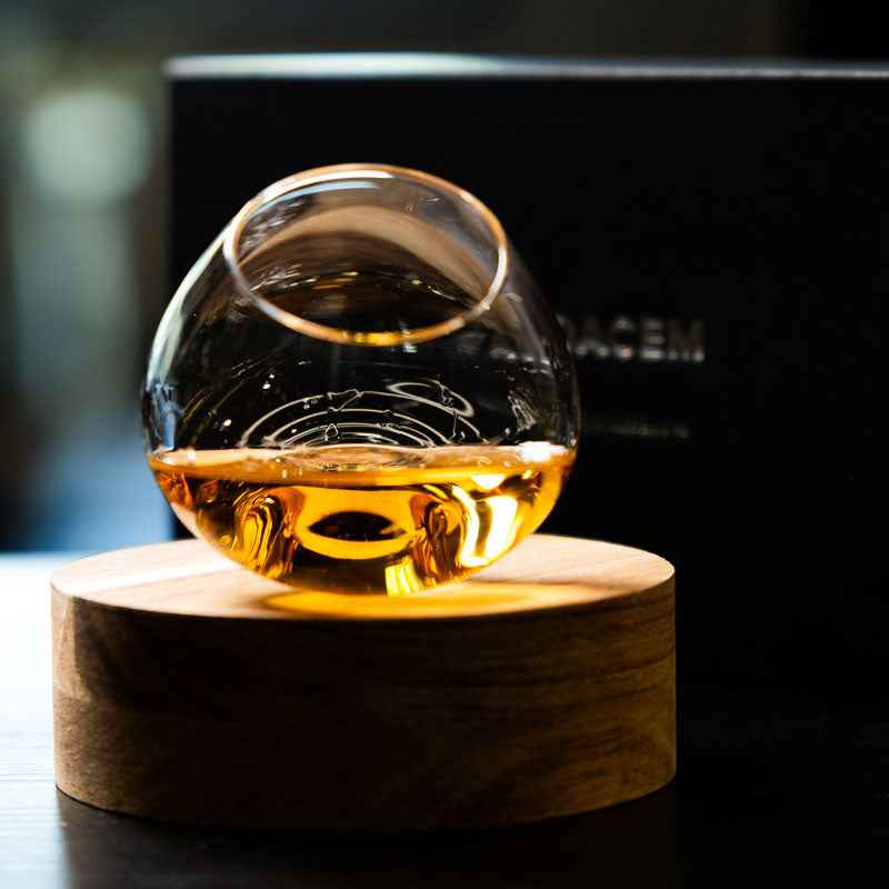 Aequilibrium Whisky Glass by Audacem filled with whisky, resting on a wooden base with the black Audacem gift box in the background
