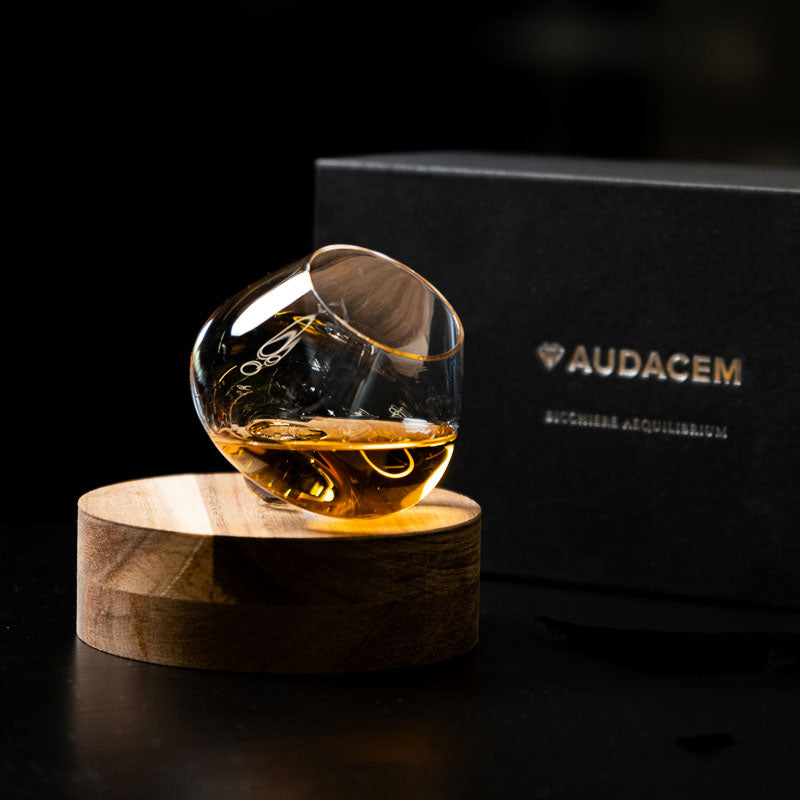 Hand-blown Aequilibrium spirit glass with whisky on wooden coaster, placed in front of black Audacem gift box.